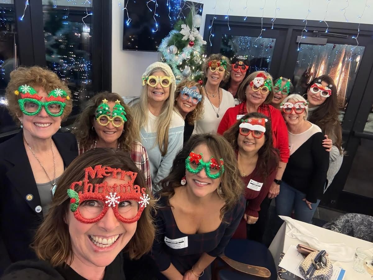 A group of smiling women at an indoor holiday party wearing festive Christmas-themed glasses and party attire.