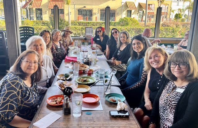 A group of people sit at a long table during an outdoor restaurant lunch, smiling and talking.