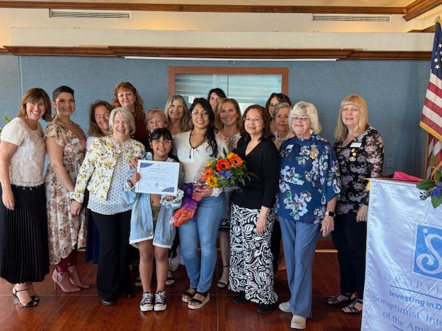 A group of people stands together indoors, with a child in the center holding an award certificate and a bouquet.