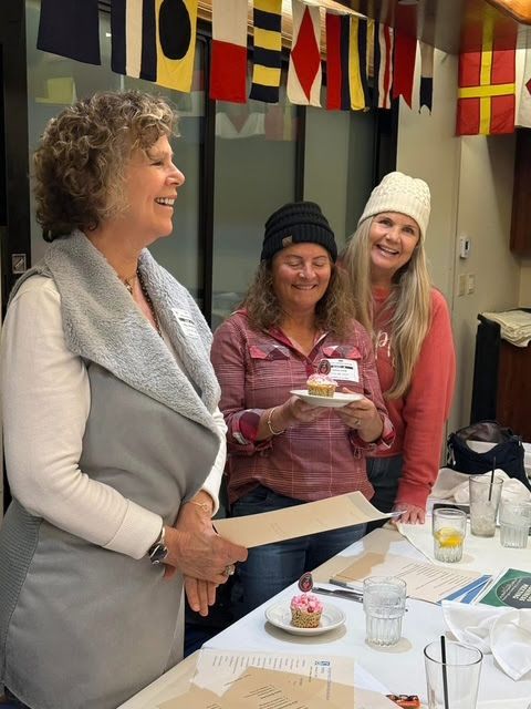 Three people smiling behind a table with cupcakes and nautical signal flags hanging in the background.