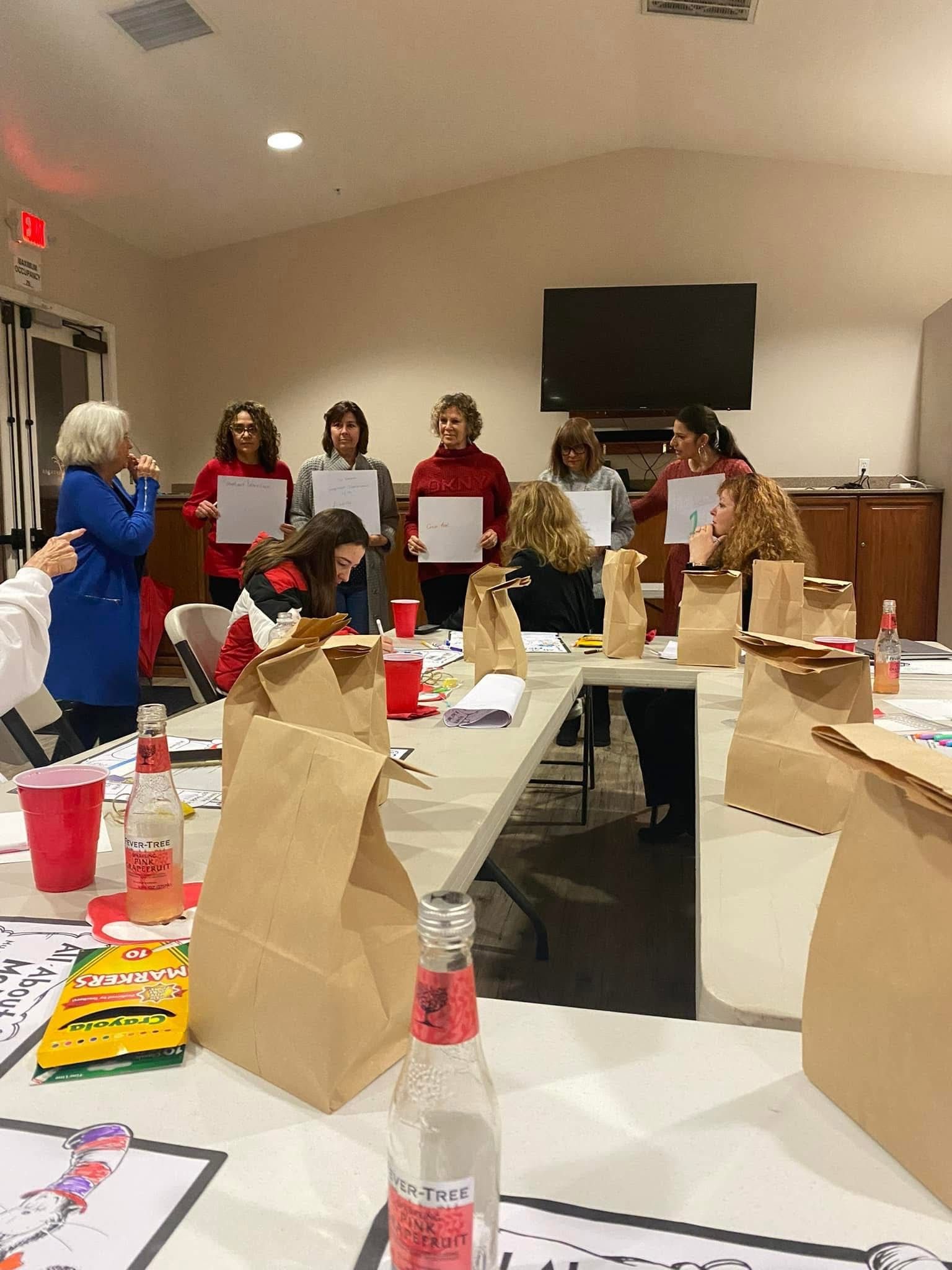 A group of people sit around a long table holding white papers in a community room with brown paper bags in the foreground.