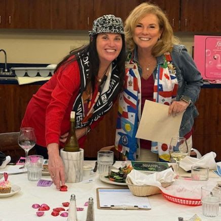 Two people smile at a table decorated with scattered rose petals, holding wine and a paper during an indoor gathering.