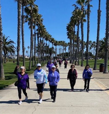 A group of people wearing purple or white shirts walk together along a paved path lined with tall palm trees.