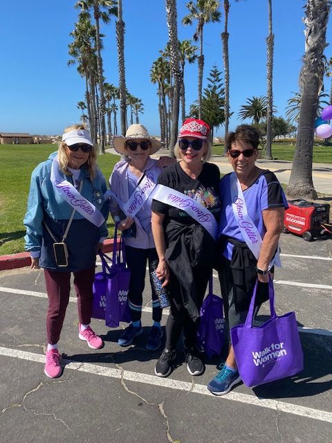Four people smiling in a sunny park wearing matching purple sashes and holding purple tote bags.