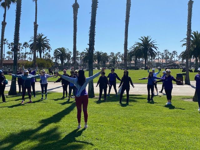 A group of people standing in a grassy park with palm trees, performing stretches with arms extended outward.