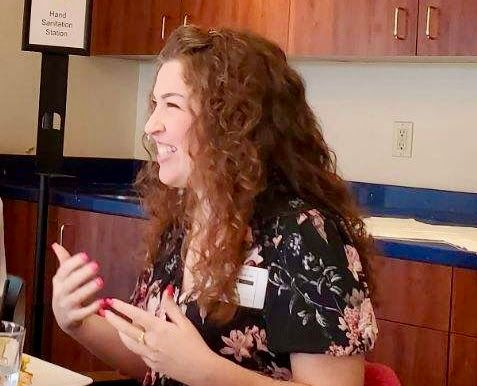 A person with long, curly brown hair and a floral top smiles while gesturing during a conversation in a room with cabinets.