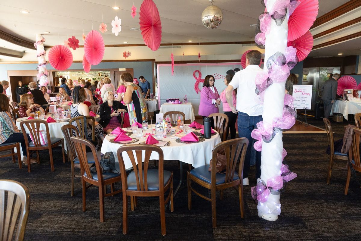 People gather at tables decorated for a pink-themed event in a banquet hall featuring pink paper fans and floral columns.