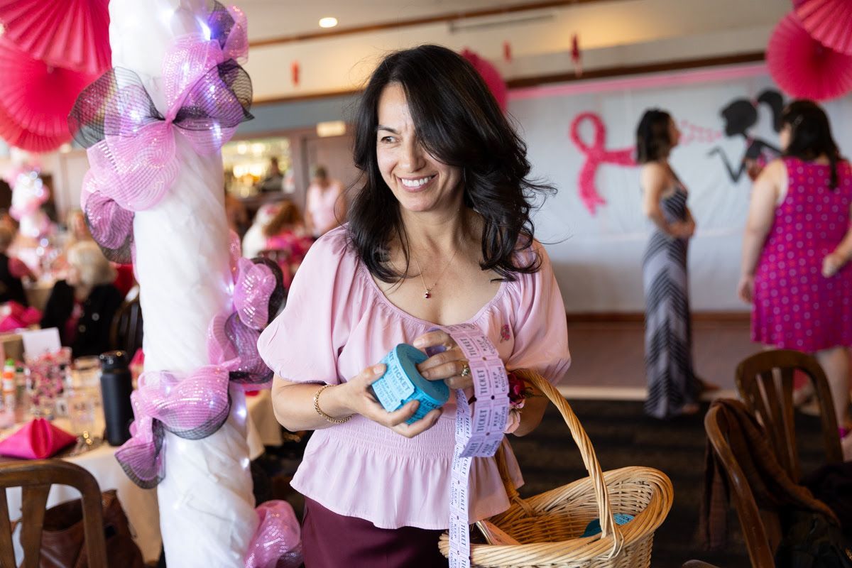 A smiling person in a pink top holds raffle tickets and a basket at a pink-themed event with a breast cancer ribbon sign.