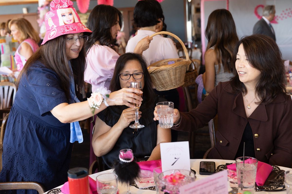 Three women toasting with drinks at a table with pink decor at a social event.