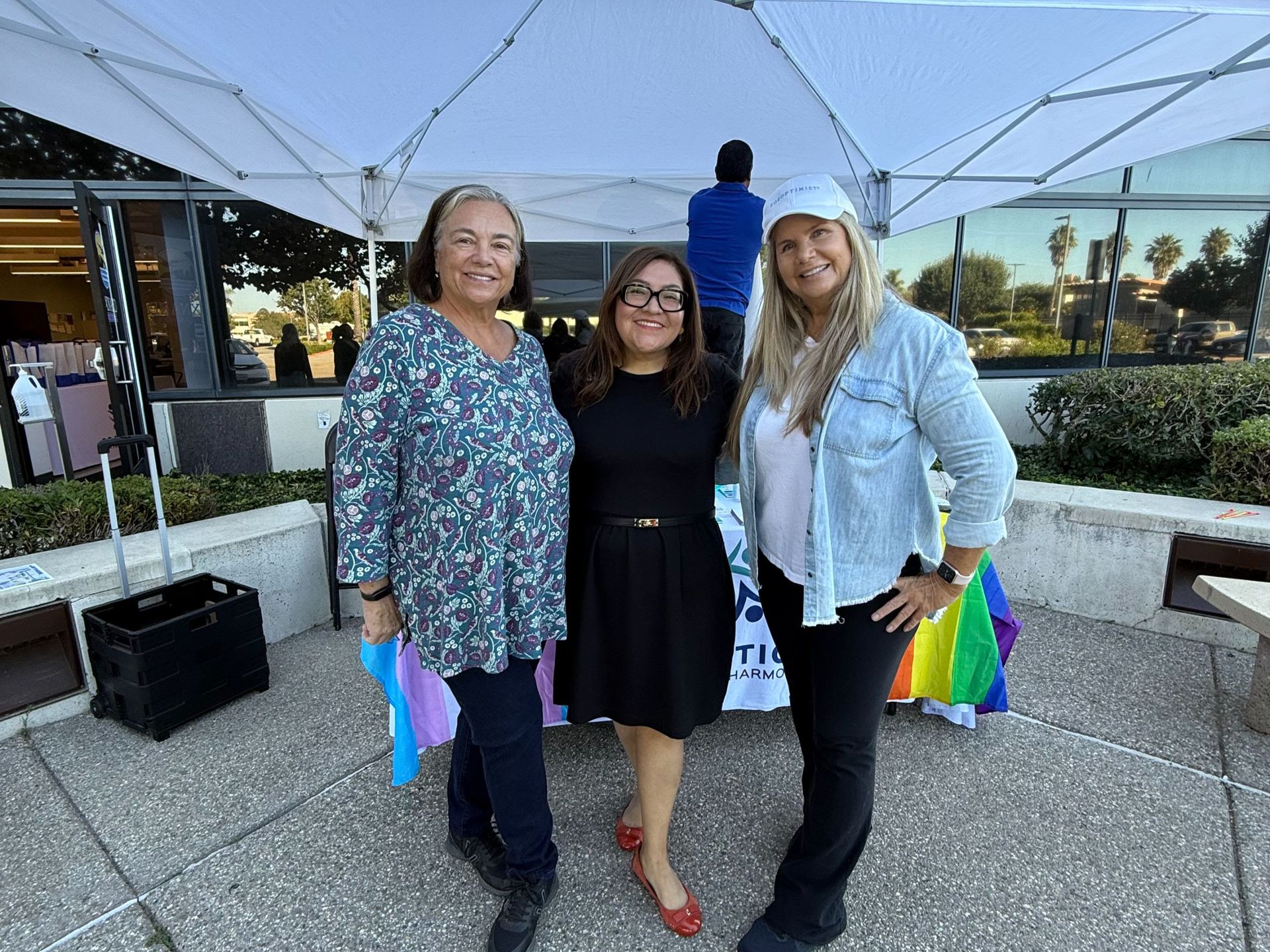 Three people smile while posing for a photo under a white tent outdoors, with Pride-colored decorations visible behind them.