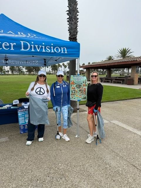 Three people standing under a Water Division tent at a park, holding litter pickers and bags during a cleanup event.
