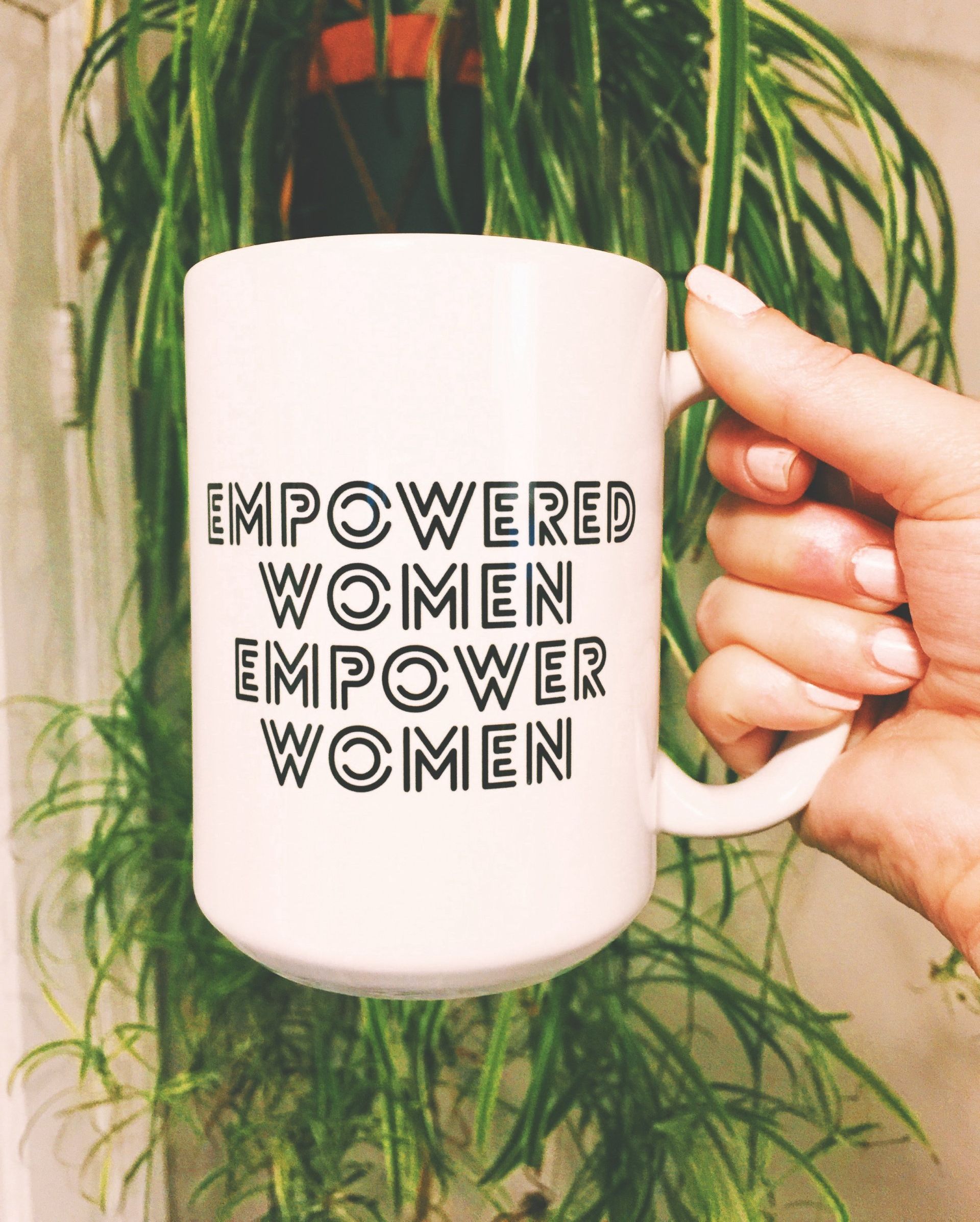 A white mug held by a hand in front of a green plant, with the text