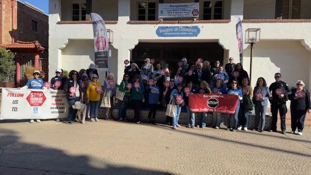 A large group of people stands in front of a building holding signs and red circular placards at an advocacy event.
