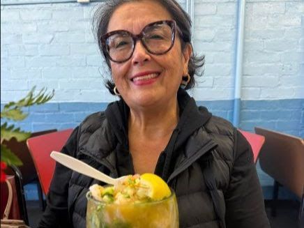 A smiling person wearing glasses and a black puffer vest holds a glass of shrimp ceviche garnished with a lemon wedge.