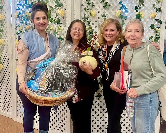 Four people smiling together, one holding a gift basket and another a small pumpkin, standing before a floral lattice wall.