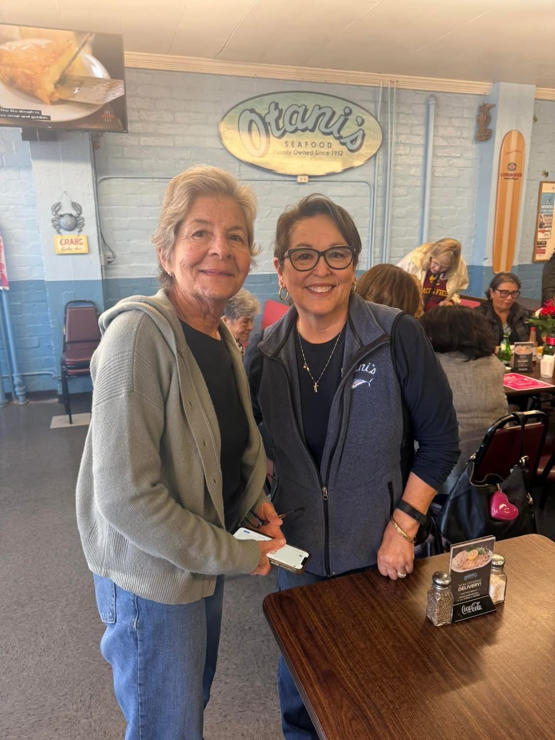 Two people stand in a restaurant, one smiling while holding a checkbook, the other wearing a vest and smiling at the camera.
