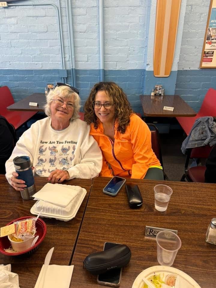 Two smiling women sit at a wooden table in a brightly lit dining area, wearing casual tops and holding various items.