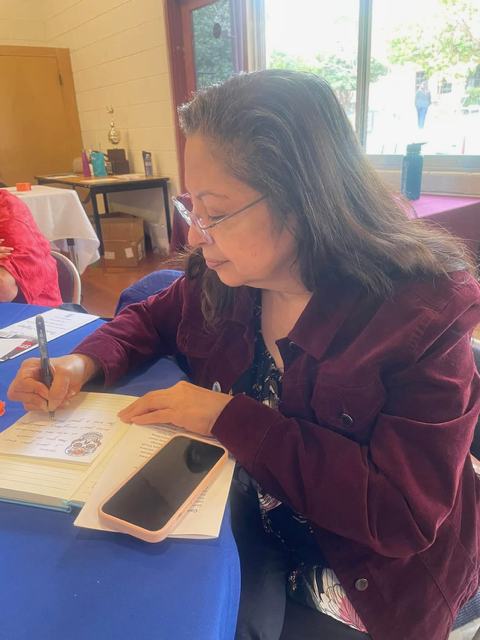 A person in a maroon jacket writes on a document at a blue table, with a smartphone placed beside the paper.