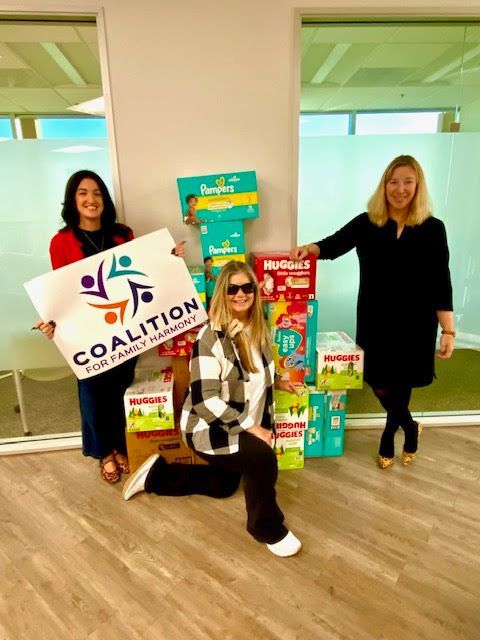 Three people stand indoors by a stack of diaper boxes; one holds a 