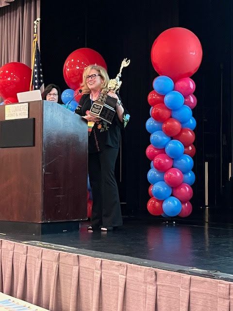 A person holding a trophy stands behind a wooden podium on a stage decorated with a tall column of red and blue balloons.