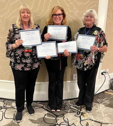 Three people standing in a row indoors holding award certificates, smiling towards the camera.