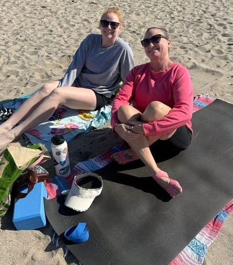 Two people wearing sunglasses sit on mats on a sandy beach, smiling toward the camera.