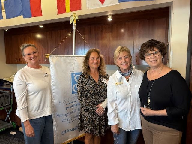 Four smiling people stand before a banner for the Soroptimist International of the Américas, indoors near wall cabinets.