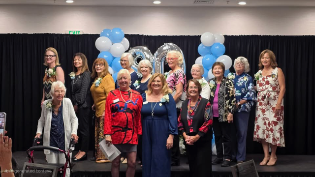 A group of people standing on a stage in front of a black curtain with blue and white balloons, celebrating together.