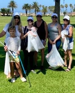 Six people stand on a grassy field in white caps, holding trash bags and a litter picker during a community cleanup event.
