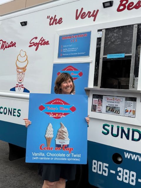 A smiling person stands in front of a Mister Softee ice cream truck, holding a sign displaying cone and cup options.