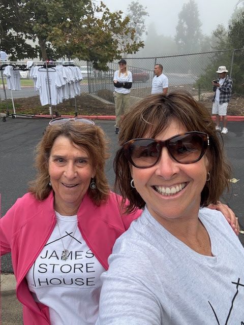 Two people smile at the camera in a parking lot, with a clothes rack and other people visible in the foggy background.
