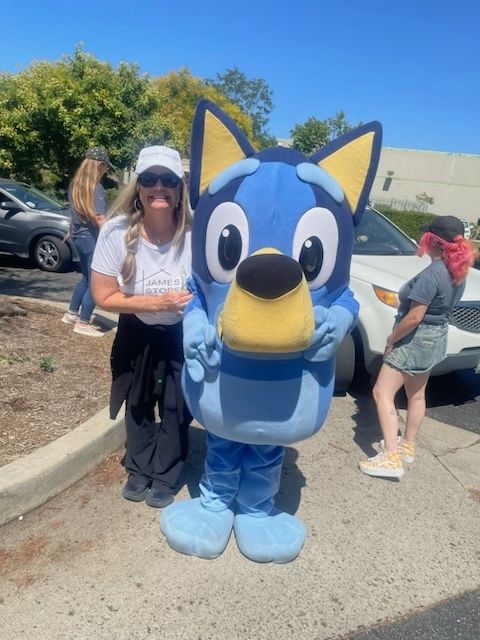 A woman in a white shirt and sunglasses poses for a photo with a Bluey character costume in an outdoor parking lot.