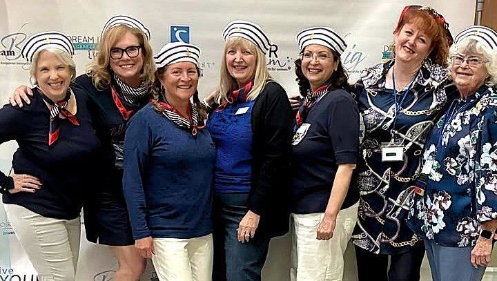 Six smiling individuals wearing striped hats and navy-themed outfits stand side-by-side in front of a white backdrop.