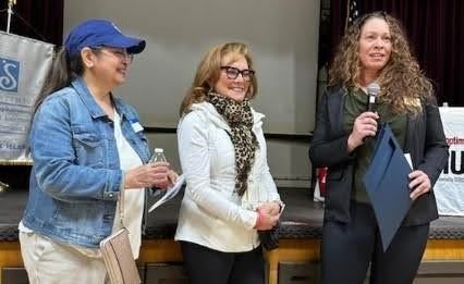 Three individuals standing on stage, one holding a microphone and folder, another smiling, in an indoor setting.