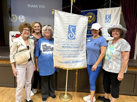 Five people stand together holding a banner for Assistance League of Channel Islands in an indoor community room.