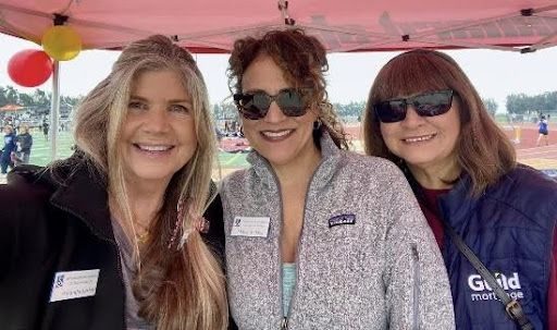 Three women wearing name tags smile together outdoors under a tent, with a blurry track and field setting in the background.
