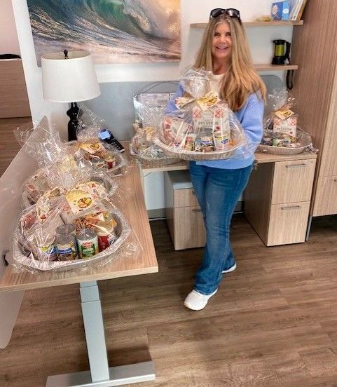 A smiling person holds a gift basket in an office filled with similar wrapped baskets on desks and tables.