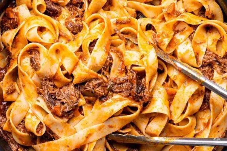 Close-up of pappardelle pasta with shredded meat sauce and serving tongs