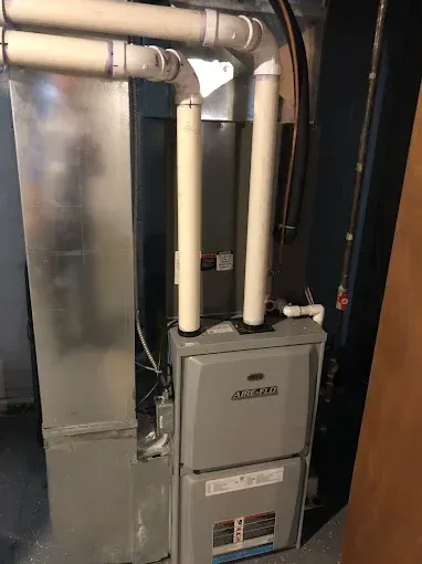 A furnace and air handler in a basement, with PVC pipes and ductwork.