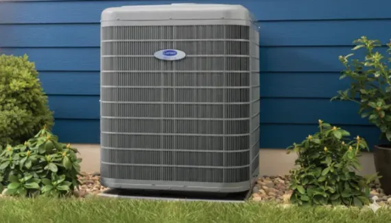 Outdoor air conditioning unit in front of a blue house with greenery and grass.