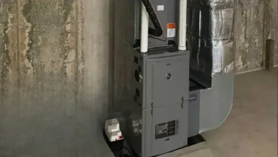 Furnace in a basement; gray metal unit, silver ductwork, next to a concrete wall.