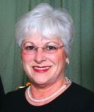Woman with short, white hair and glasses smiles; wearing a black top and pearl necklace.