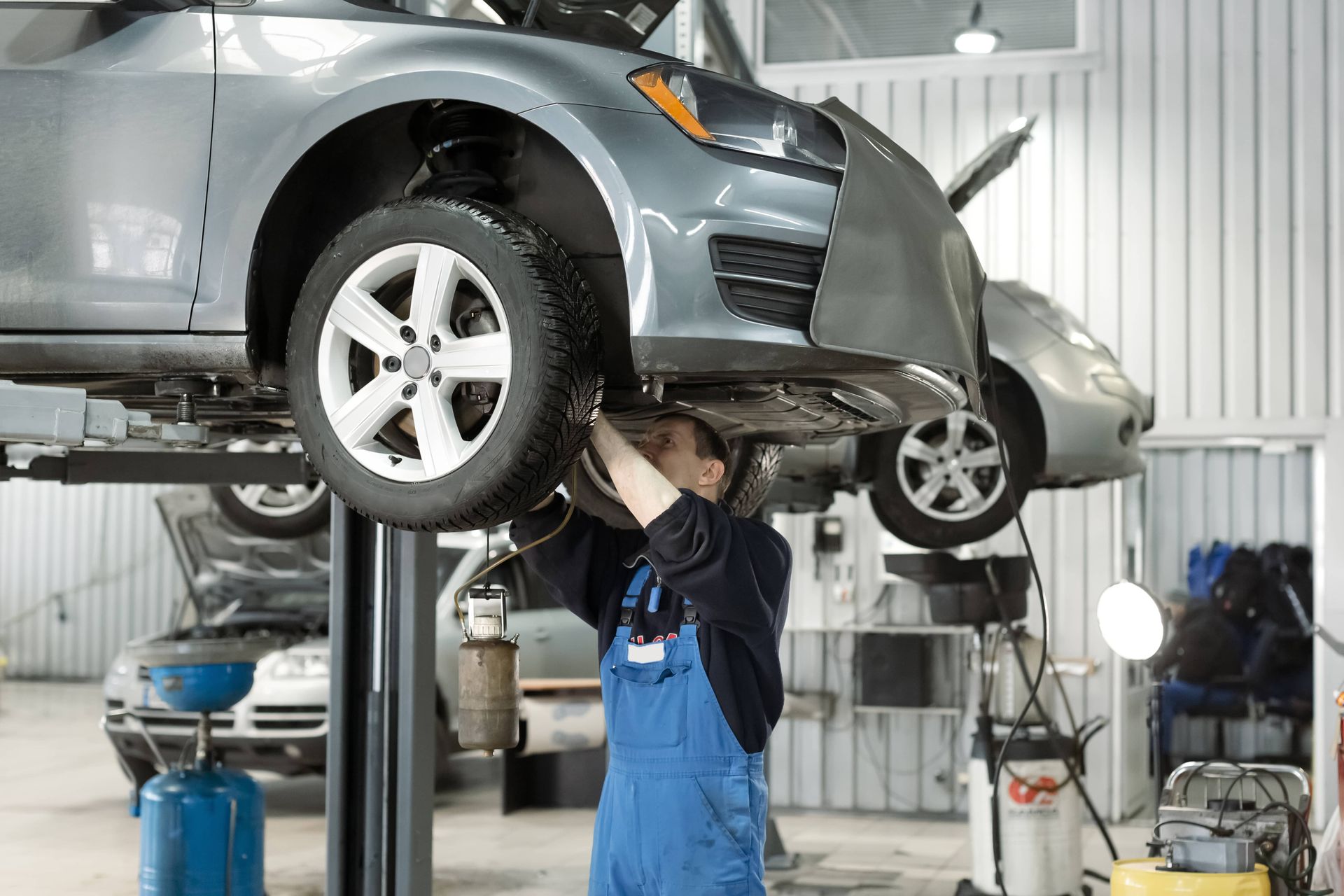 Mechanic in blue overalls working on a gray car raised on a lift in a garage.