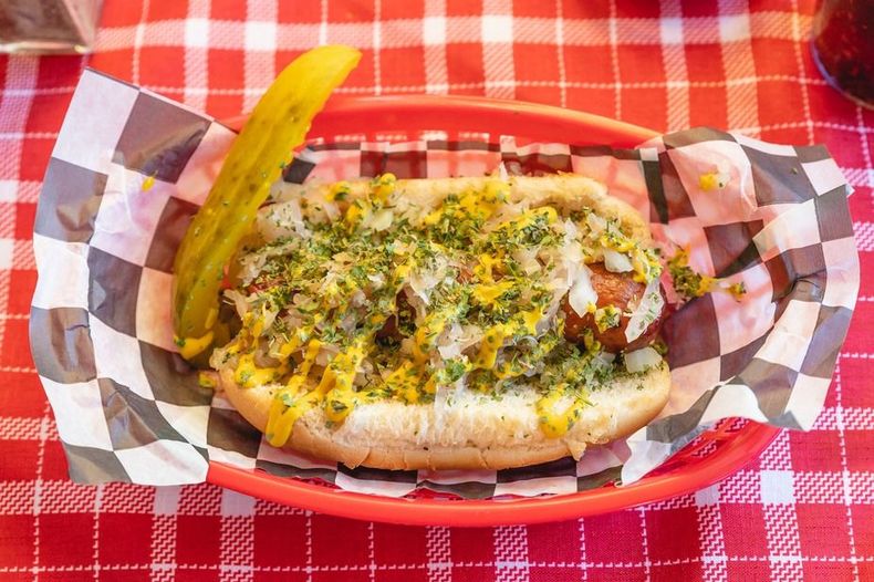 Grilled hot dog with mustard, herbs, and a pickle in a red basket on a red checkered tablecloth