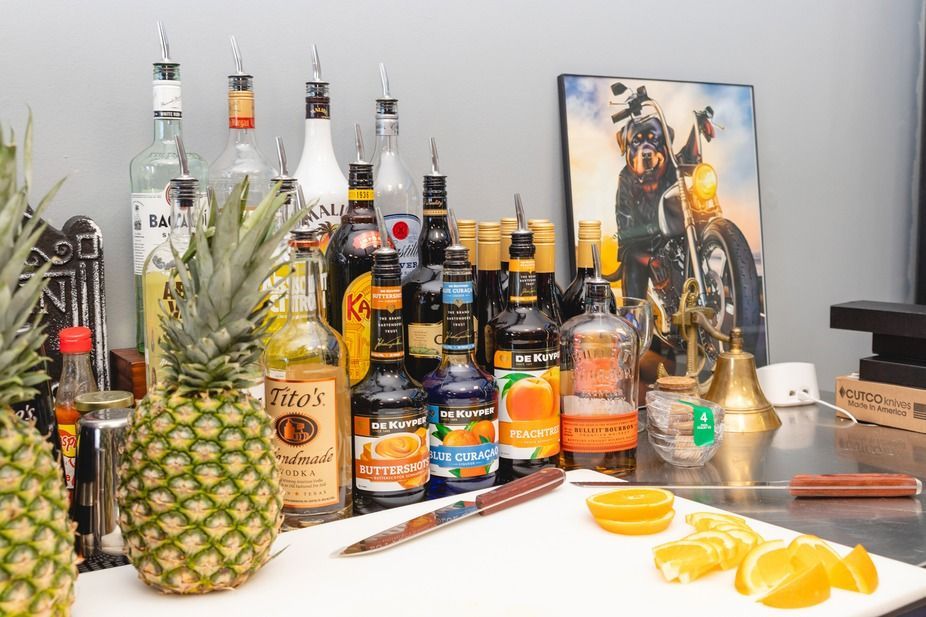 Tropical drink setup with liquor bottles, pineapples, citrus slices, and a dog painting on a table