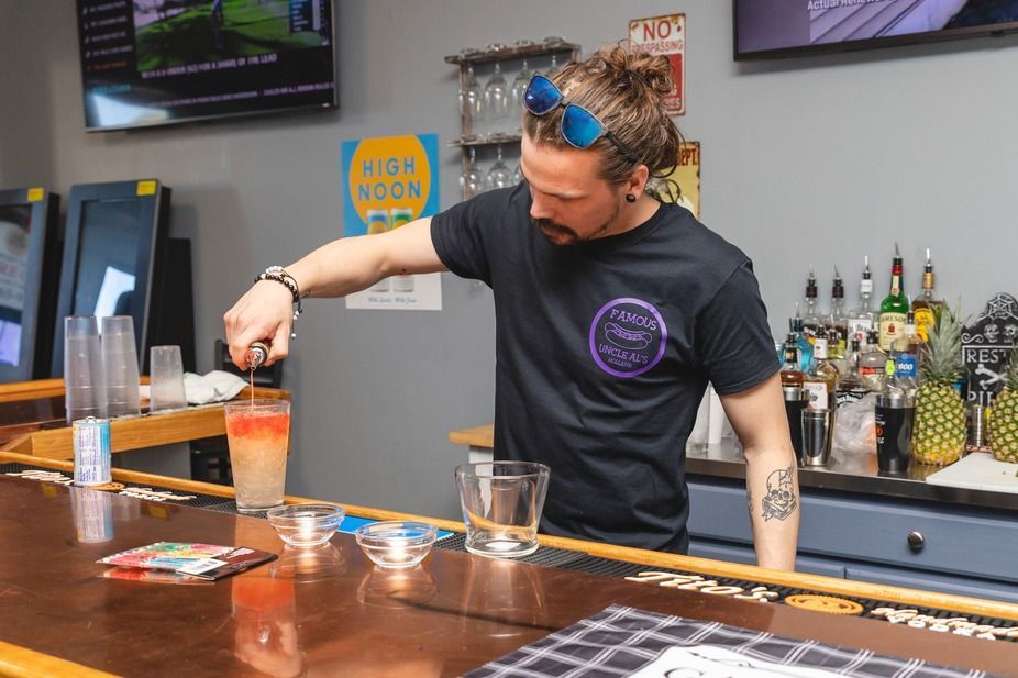 Bartender stirring an orange cocktail behind a bar with bottles and a TV in the background
