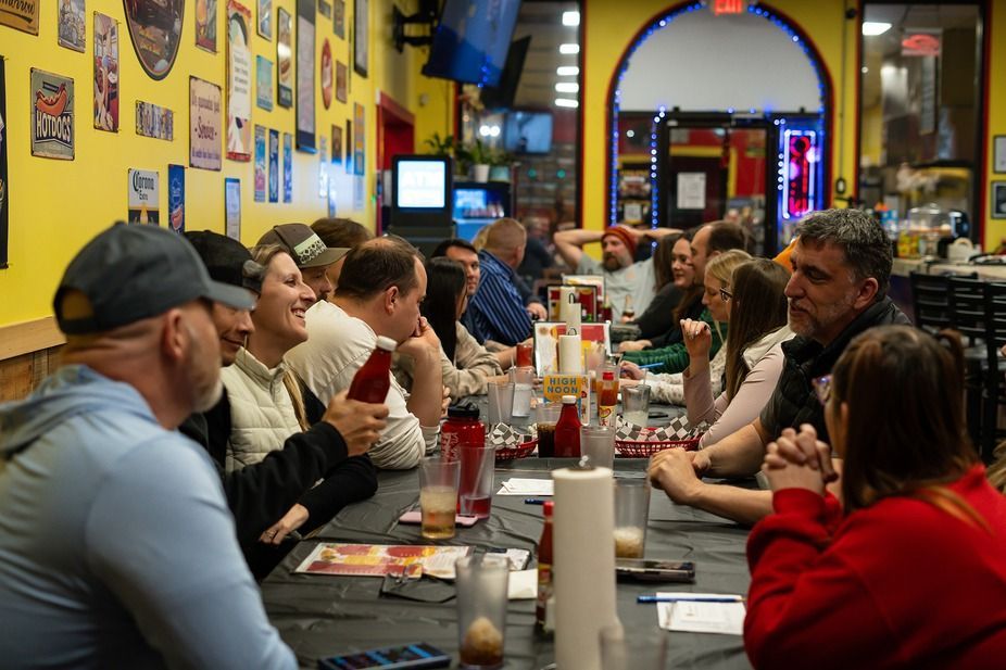 People seated at a long table in a casual restaurant, eating and talking under warm lights.