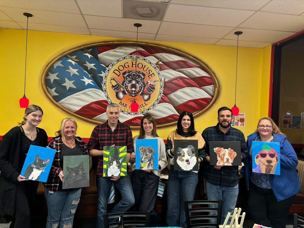 Six people hold colorful cat portraits in front of a large mural with an American flag and school emblem.