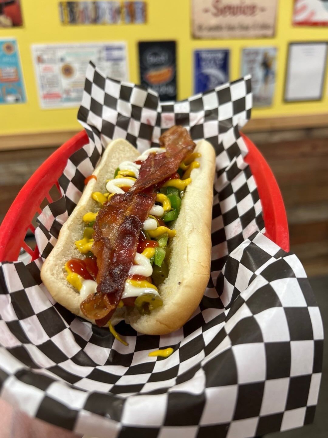 Hot dog topped with bacon, mustard, and relish in a red basket with black-and-white checkered paper
