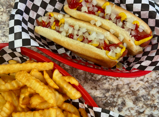 Two hot dogs with mustard and onions in a basket, with crinkle-cut fries on a table nearby